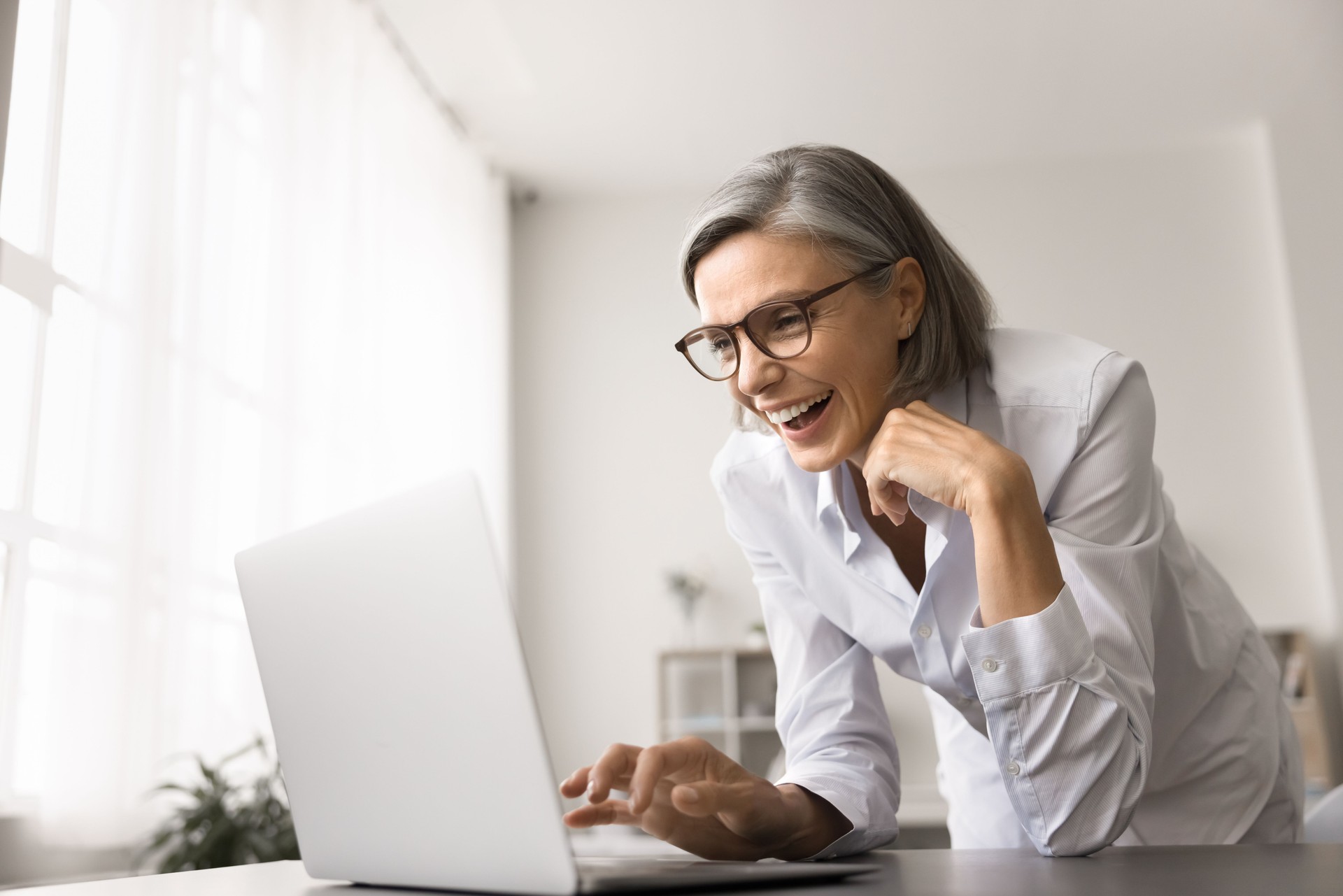 Laughing mature businesswoman with glasses leaned at desk using laptop